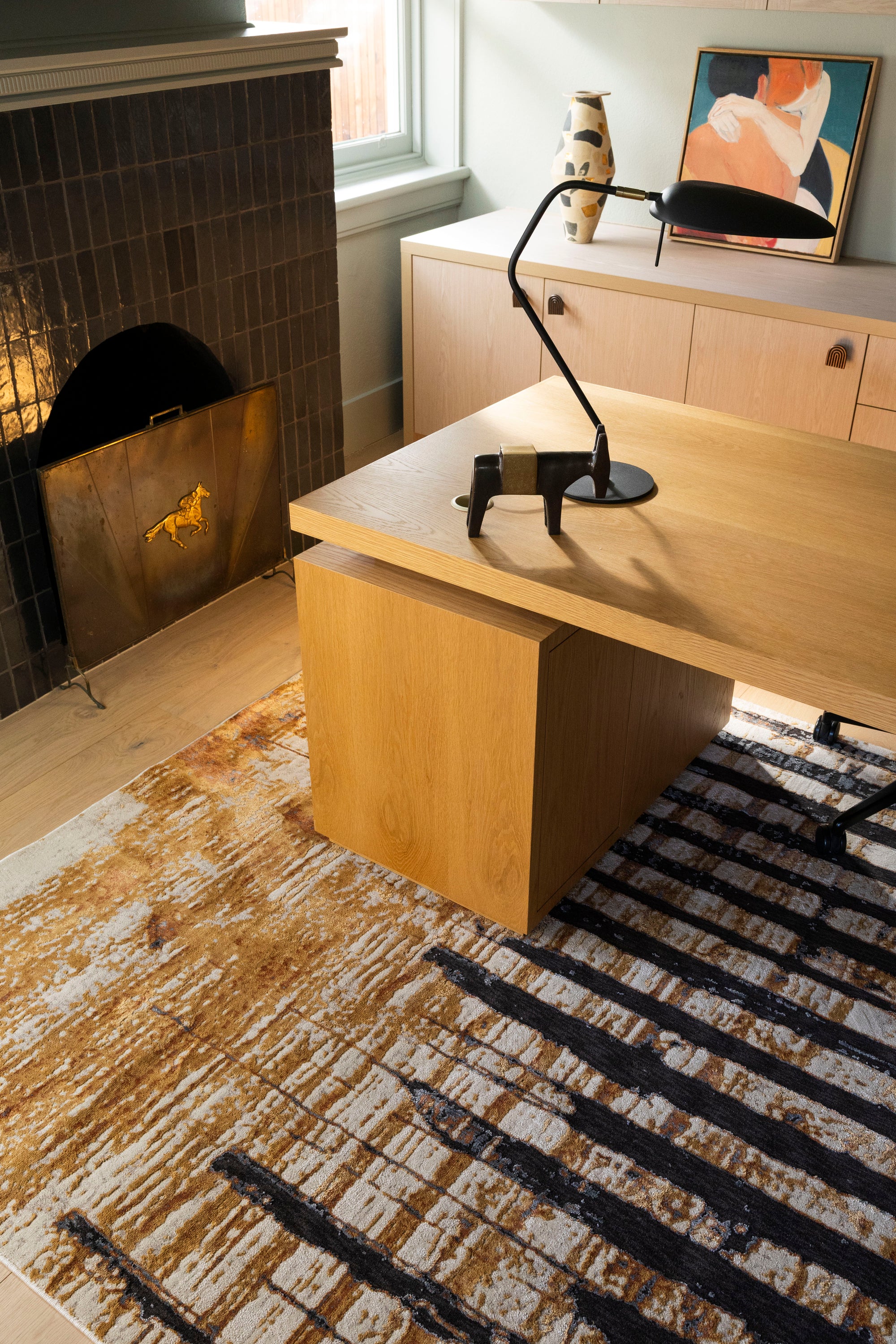 Home office wooden desk with a lamp in a room with a fireplace and Amber Smoke Abstract Rug by Four Corners Rugs