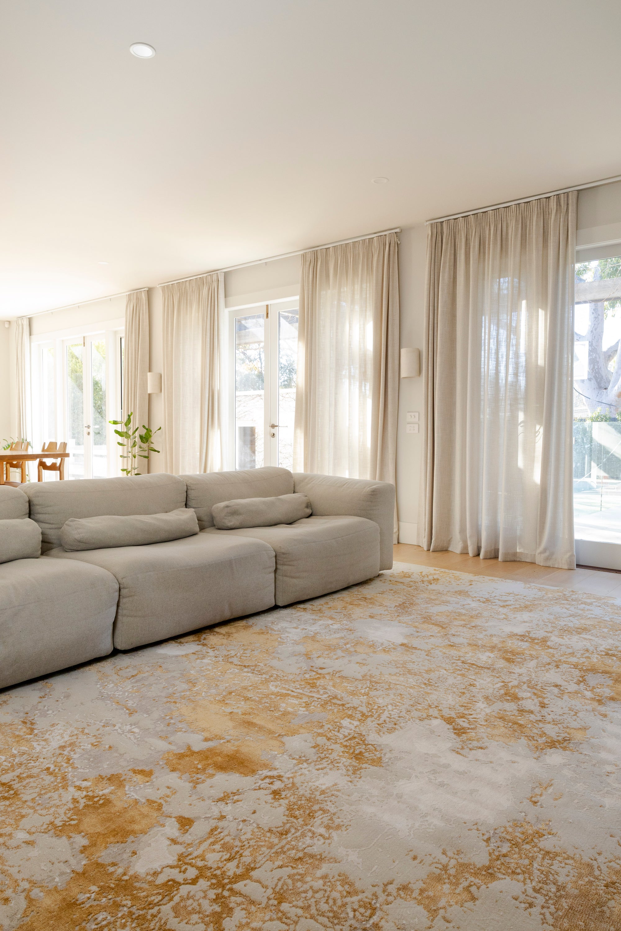 Modern living room with a grey sofa, Golden Dusk Abstract Rug by Four Corners Rugs, and large windows with white curtains.