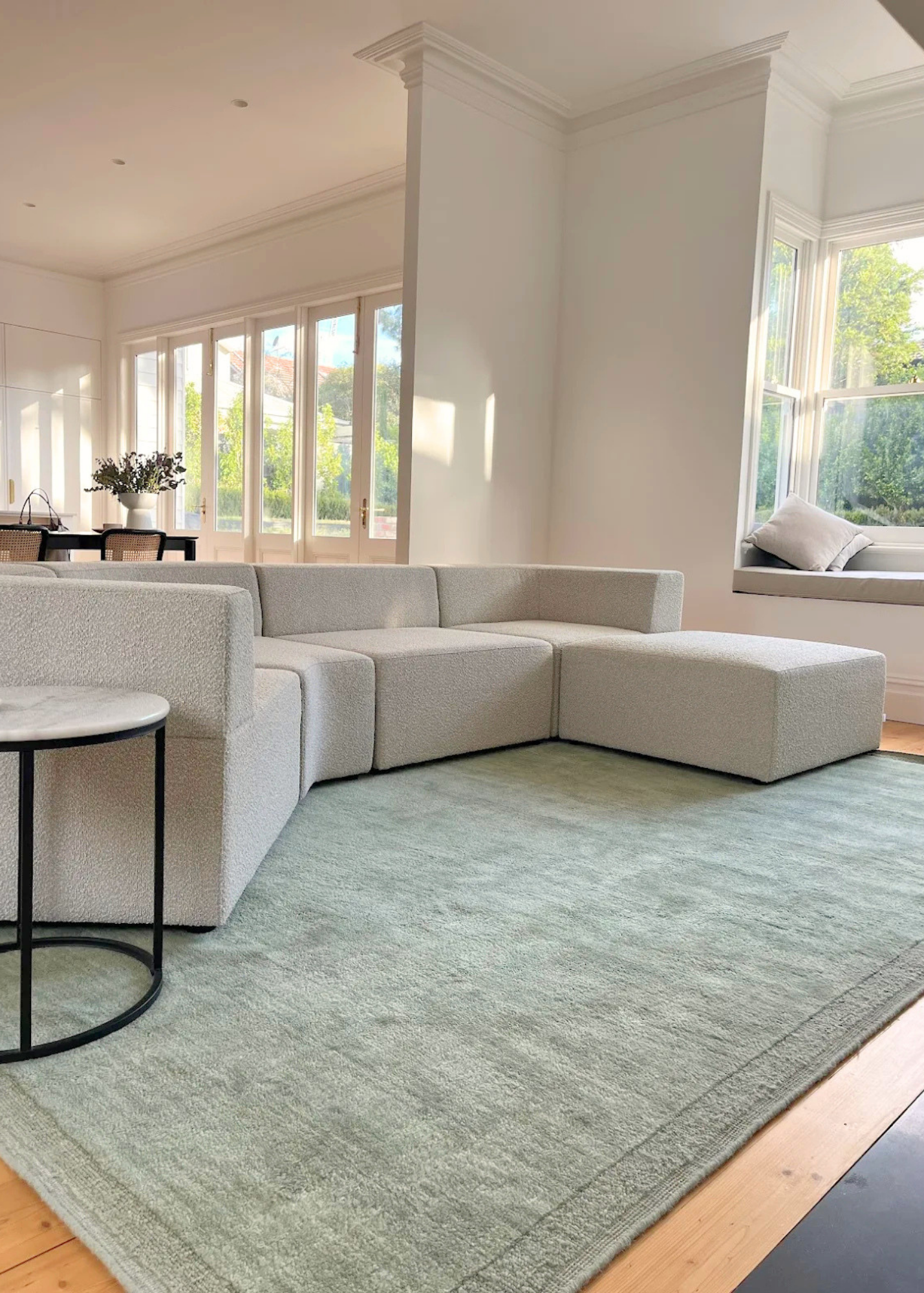 Modern living room with a light grey sectional sofa, Double Border Mint Wool Rug by Four Corners Rugs, and large windows.