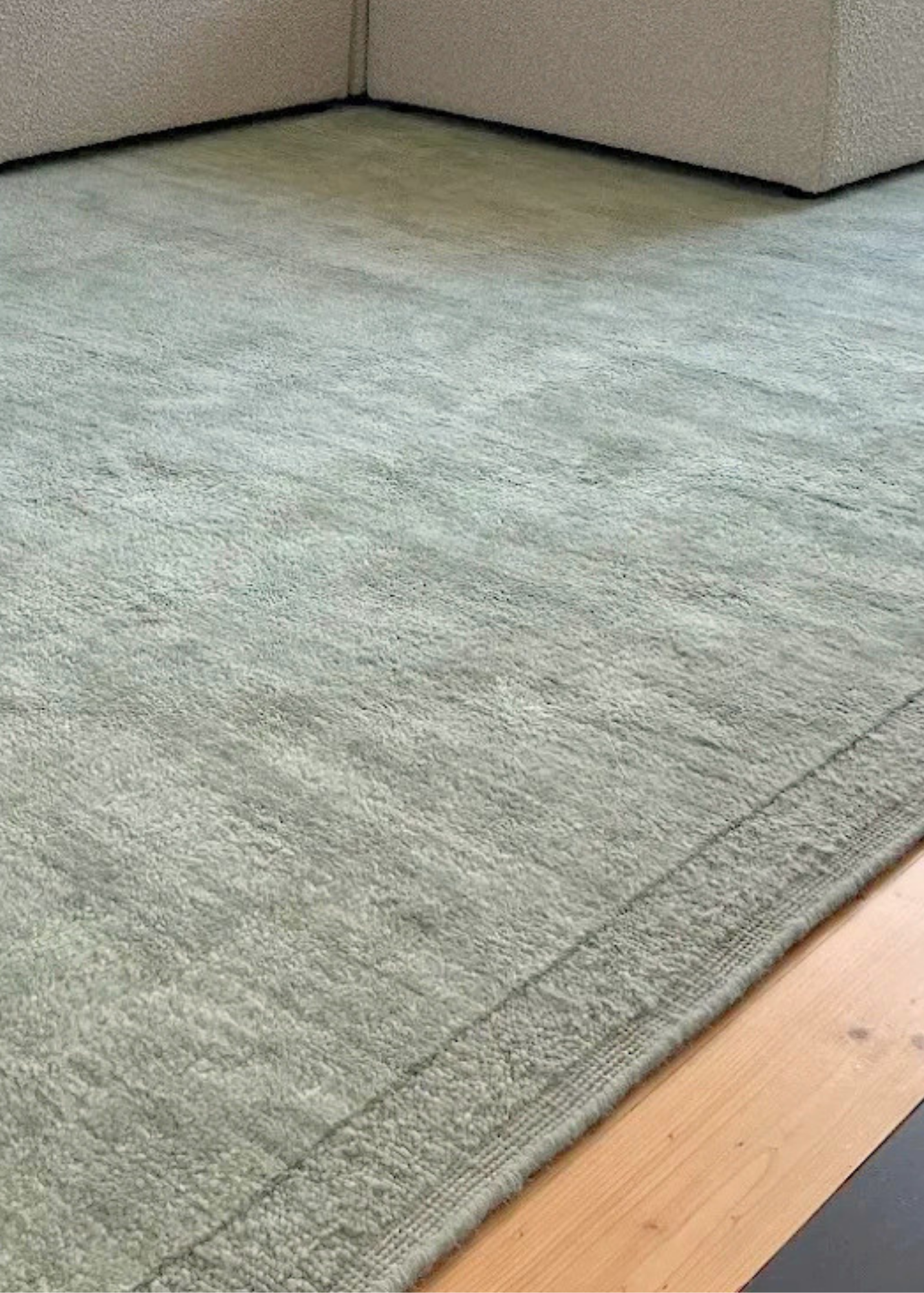 Double Border Mint Wool Rug by Four Corners Rugs on a wooden floor with a corner of a beige sofa visible.